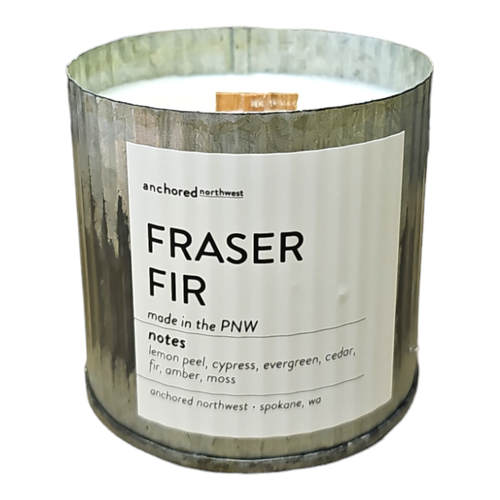 The Anchored Northwest Fraser Fir Rustic Vintage Farmhouse Wood Wick Candle features a soy wax blend and comes in a charming rustic metal holder. The label highlights fragrance notes of lemon peel, cypress, evergreen, cedar, fir, amber, and moss. Created by Anchored Northwest in Spokane, WA.