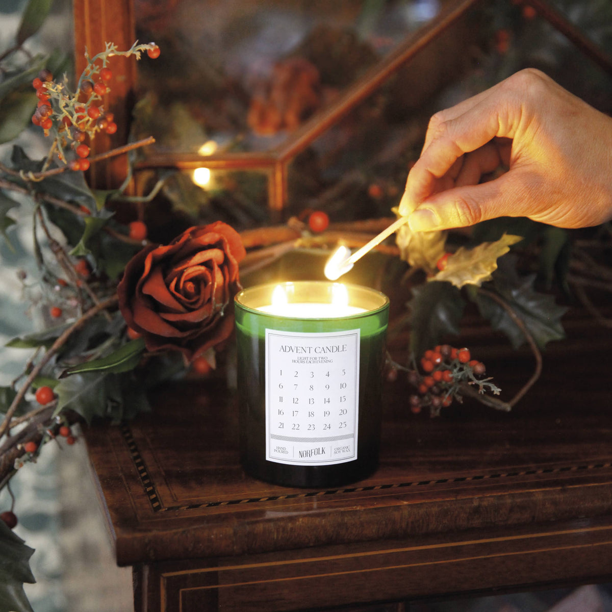 A hand lights the Norfolk Natural Living Advent Christmas Candle by Norfolk Natural Living on a wooden table adorned with red berries, holly leaves, and a red rose, creating a festive holiday atmosphere.