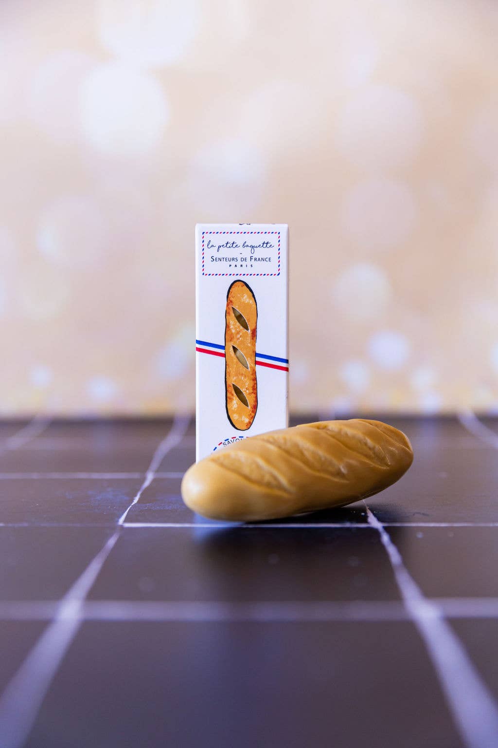 The "Baguette" Soap by Senteurs De France rests on dark tiles before its white box with a baguette-shaped window and French-themed design, capturing the cozy charm of a Parisian bakery in warm light.