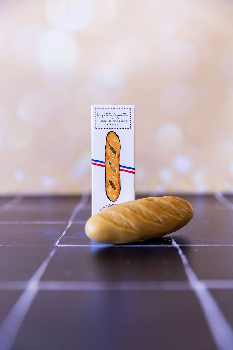 The "Baguette" Soap by Senteurs De France rests on dark tiles before its white box with a baguette-shaped window and French-themed design, capturing the cozy charm of a Parisian bakery in warm light.
