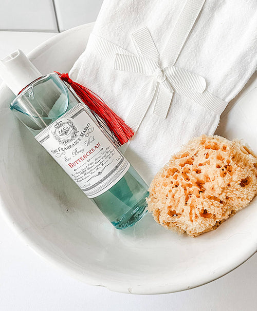 A bottle of Buttercream-scented fragrance mist from Z&Co.'s Farmhouse Collection, featuring a red tassel, is elegantly placed in a white bowl beside a natural sea sponge and folded white cloth tied with a ribbon, all resting on a light surface.