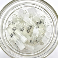 A glass jar filled with Perfectly Scentsable Perfume Oil Samples from the brand Perfectly Scentsable. The small, clear, plastic vials with white caps and labels seem to be sample containers for perfumes or essential oils and are arranged haphazardly within the jar. The bright lighting accentuates their transparency.