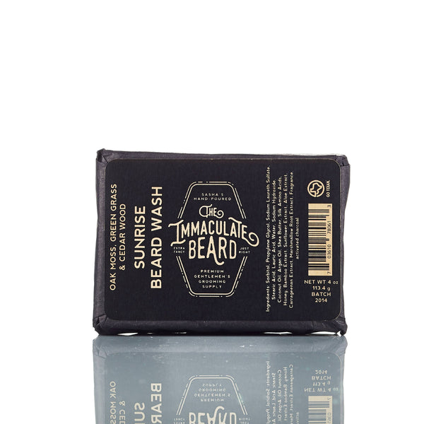 The Immaculate Beard SUNRISE Beard Wash Bar, in black packaging with white and gold text, stands on a reflective surface. Its oak moss, green grass, and cedarwood scent makes it perfect for daily beard care.