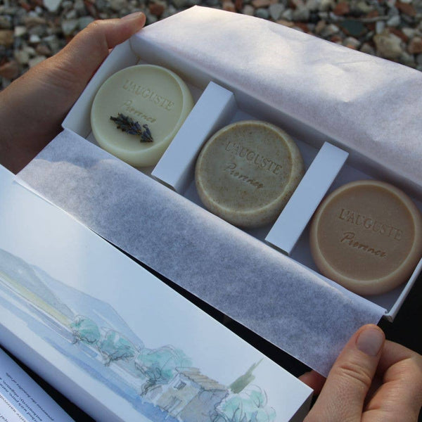 Hands hold an open tissue-lined box containing three round, handcrafted soaps engraved with "L'AUGUSTE Provence." Below the set, a watercolor landscape illustration evokes Provencal olive pit powder hues.
