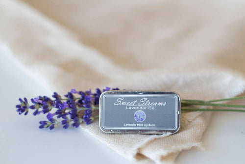 A Sweet Streams Lavender Co. Lavender & Mint Lip Balm metal tin sits on a beige cloth with a lavender sprig. Designed with natural ingredients, the gray tin showcases a purple lavender logo and text, emphasizing its calming lavender peppermint essence.