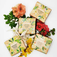 Four floral-themed soap bars, including Senteurs De France’s organic ylang ylang essential oil soap, are displayed with fresh flowers and leaves. Each bar features botanical illustrations and elegant text on decorative packaging.