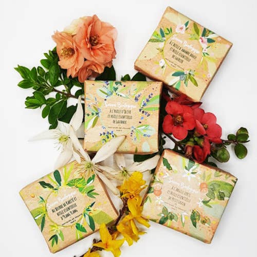 Four floral-themed soap bars, including Senteurs De France’s organic ylang ylang essential oil soap, are displayed with fresh flowers and leaves. Each bar features botanical illustrations and elegant text on decorative packaging.