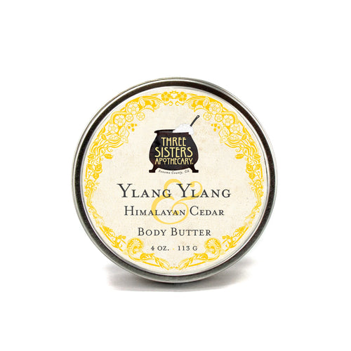 A 4 oz round tin of Three Sisters Apothecary Ylang-Ylang Body Butter features a yellow floral-bordered label—ideal for anyone seeking natural, hydrating skincare.