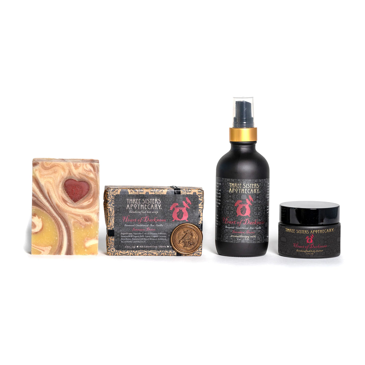 A collection of personal care products from Three Sisters Apothecary features the Heart of Darkness Intensions Bar Soap, a soap box with a coin, a spray bottle with Rosewood oil, and a jar containing essential oils. Each item is elegantly adorned with the brand’s logo.