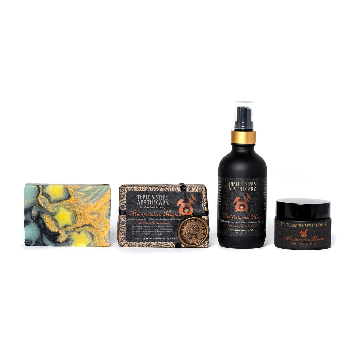 A set of Three Sisters Apothecary products is displayed against a white background. The set includes a swirled soap bar named "Intensions Bar Soap - Manifestation Magic," a rectangular soap with a round emblem, a spray bottle, and a small jar. All items feature intricate, dark-colored labels with an ornate logo embodying ancestral wisdom and text.