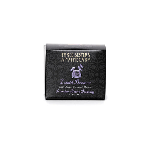 A black rectangular box labeled "Three Sisters Apothecary" contains a product named "Intentions Body Butter - Lucid Dreaming" from their Intentions line. The label features a dark floral pattern and lists ingredients such as Balsam, Precious Cedarwood, Wormwood, and Mugwort, claiming to aid in "Intense Active Dreaming." 1.7 oz (48 g).