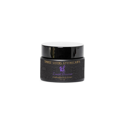 A small jar labeled "Three Sisters Apothecary Intentions Body Butter - Lucid Dreaming" in purple text. The jar is black with a dark, intricate pattern and has a matching black lid, featuring hints of precious cedarwood for an elevated sensory experience.