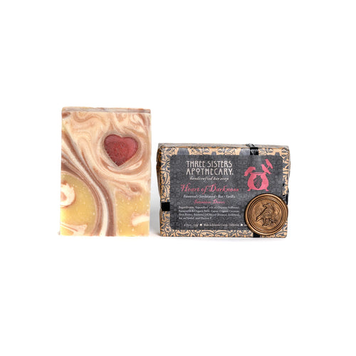 Two bars of soap are shown. Both belong to the Three Sisters Apothecary brand. The bar on the left is called Heart of Darkness and features a marbled design with a red heart embedded in the top. The bar on the right is elegantly wrapped in decorative paper, labeled "Three Sisters Apothecary," and includes a wax seal, exuding hints of essential oils.