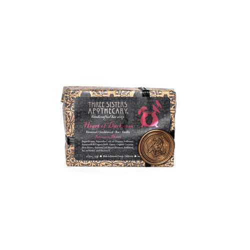 A bar of Three Sisters Apothecary Intensions Bar Soap - Heart of Darkness, elegantly wrapped in decorative paper and adorned with a gold wax seal that showcases an embossed figure, possibly depicting a bird or mythical creature. This luxurious soap is infused with essential oils, enhancing its allure with dark design elements on the label.