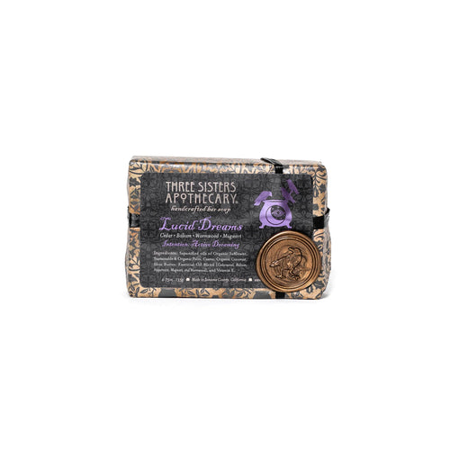 A bar of soap labeled "Three Sisters Apothecary Intensions Bar Soap - Lucid Dreaming" is shown. The wrapper features a dark, intricate design with the product name "Lucid Dreams" in the center. A round emblem with a mermaid illustration is affixed to the bottom right corner, evoking an aura of deep sleep and enhancing psychic abilities.