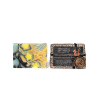 Two handcrafted soap bars are displayed. The one on the left features a swirling pattern of green, yellow, and black. The one on the right is partially wrapped in detailed packaging labeled "Three Sisters Apothecary" with a wax seal and a "Manifestation Magic" tagline inspired by ancestral wisdom, identified as the Three Sisters Apothecary Intensions Bar Soap - Manifestation Magic.