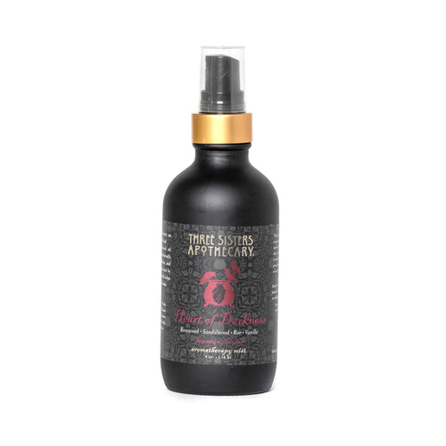 A black bottle of "Three Sisters Apothecary Intentions Aromatherapy Mist - Heart of Darkness" features a gold cap and a spray nozzle. The label includes a red rose graphic and decorative designs with text details about the compassionate essential oils in the Heart of Darkness blend.