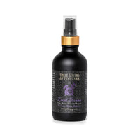 A bottle of "Three Sisters Apothecary Intentions Aromatherapy Mist - Lucid Dreaming" is shown. The bottle is matte black with a gold top and sprayer. The label features a purple design and text indicating it is a sleep-aid product containing essential oils, including soothing Cedarwood and other botanicals.