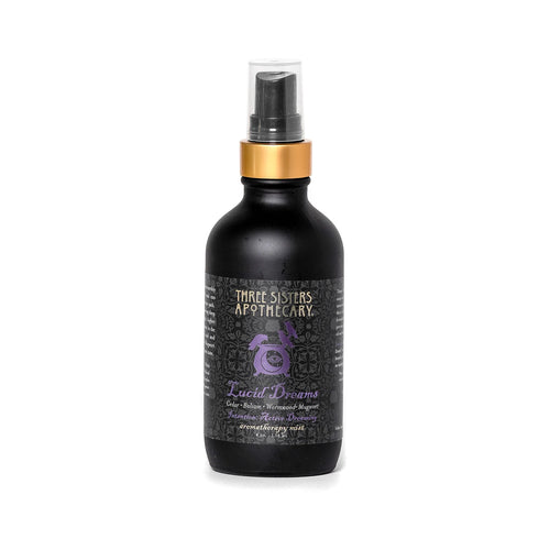 A bottle of "Three Sisters Apothecary Intentions Aromatherapy Mist - Lucid Dreaming" is shown. The bottle is matte black with a gold top and sprayer. The label features a purple design and text indicating it is a sleep-aid product containing essential oils, including soothing Cedarwood and other botanicals.