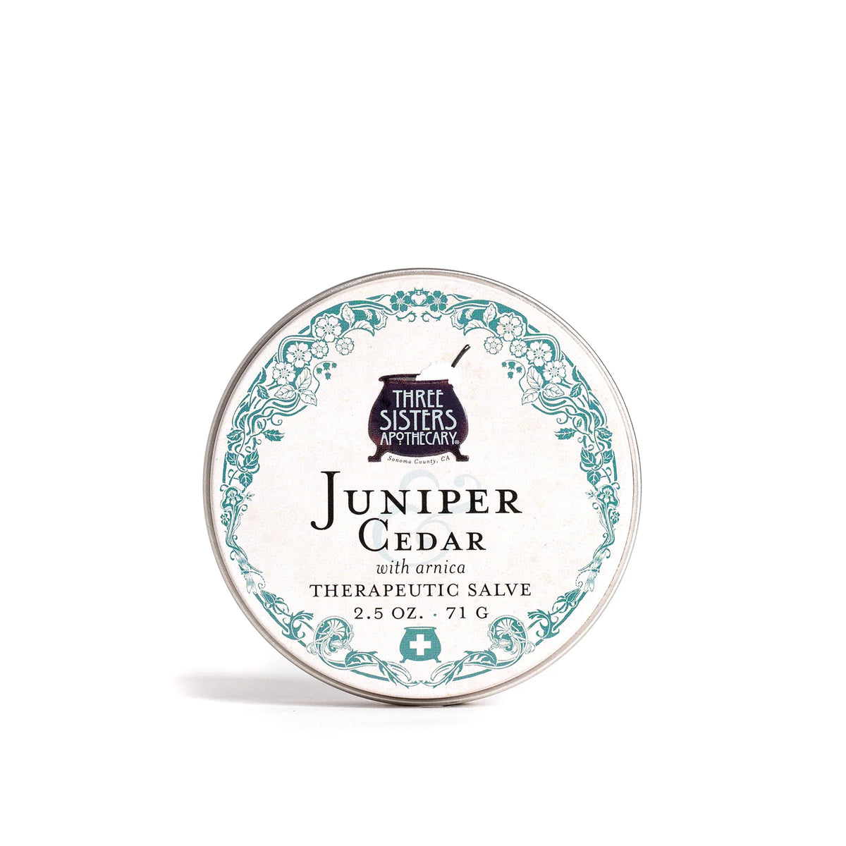 A round tin labeled "Three Sisters Apothecary Juniper & Cedar Salve" features teal floral accents on white—an herbal blend designed for soothing sore muscles and supporting natural skin restoration.
