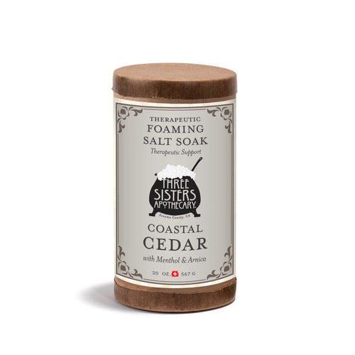Three Sisters Apothecary Therapeutic Foaming Coastal Cedar (20oz) features menthol and arnica to help relieve muscle tension. The cylindrical container has a gray label with decorative borders and a small cauldron illustration.