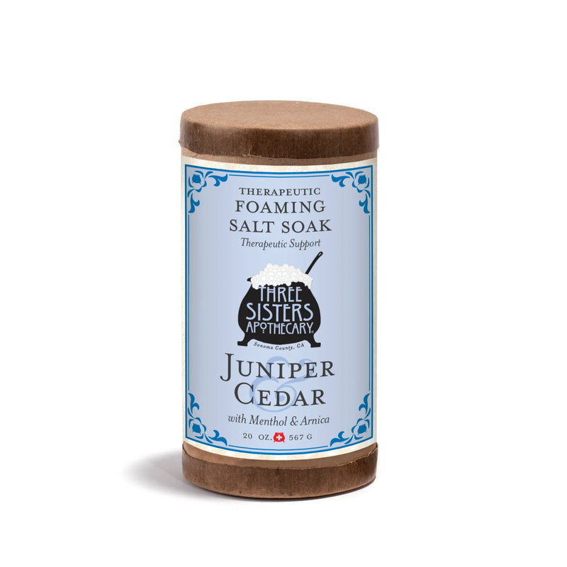 Three Sisters Apothecary Therapeutic Foaming Juniper Cedar - 20oz bath soak comes in a cylindrical container with a blue, white, and brown label. Infused with Pacific sea salts, it's designed for muscle relief.