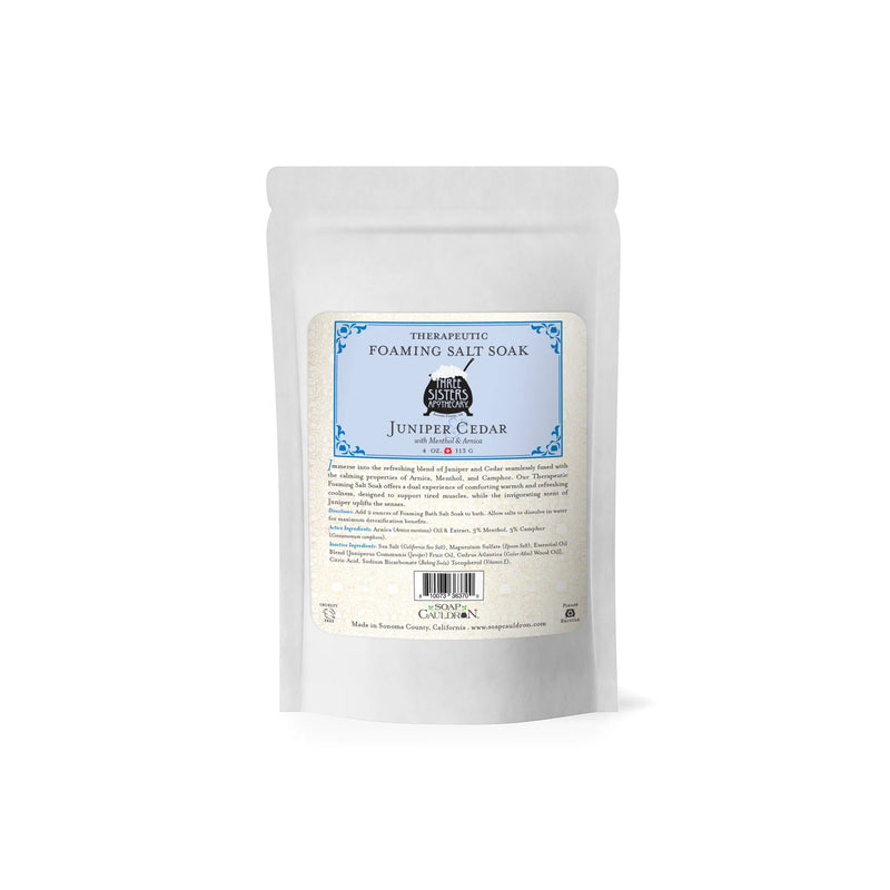 A white, resealable pouch labeled "Three Sisters Apothecary Therapeutic Foaming Juniper Cedar - 20oz" features blue accents and product details. Infused with Pacific sea salts, it offers a relaxing muscle relief bath experience. Brand: Three Sisters Apothecary.