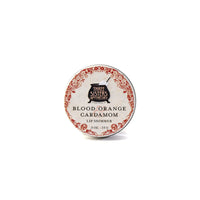 A round tin of hydrating Three Sisters Apothecary Blood Orange & Cardamon Lip Shimmer, made with natural beeswax, has a vintage-style red and white label with black text.