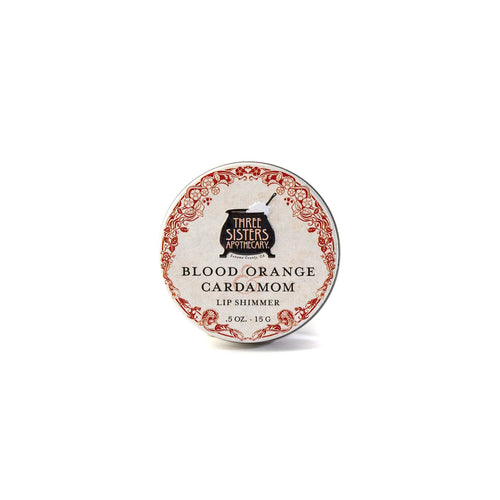 A round tin of hydrating Three Sisters Apothecary Blood Orange & Cardamon Lip Shimmer, made with natural beeswax, has a vintage-style red and white label with black text.