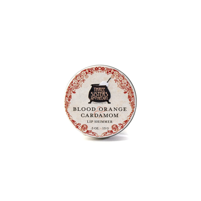 A round tin of hydrating Three Sisters Apothecary Blood Orange & Cardamon Lip Shimmer, made with natural beeswax, has a vintage-style red and white label with black text.
