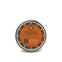 A round tin with a metal lid displaying the label "Three Sisters Apothecary Shave Balm - Bay Rum" in orange and black. The label features a beard icon, and the scent is "Bay Rum." Infused with therapeutic plant oils, the 2.5 oz (71 g) balm offers immediate relief for post-shave skin.