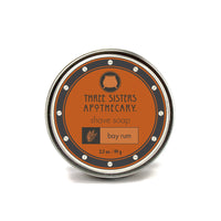 This circular tin container of Three Sisters Apothecary Shave Soap in the Bay Rum scent features an eye-catching label with an orange and black design, topped with a cauldron icon. The 3.5 oz / 99 g shave soap is enriched with essential oils, providing excellent moisture and nourishment.