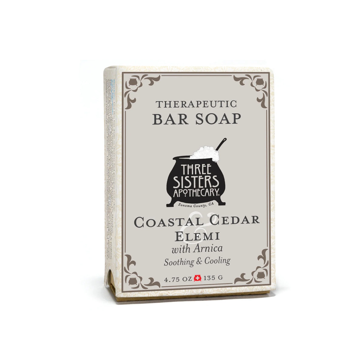 A 4.75 oz (135 g) Three Sisters Apothecary Coastal Cedar & Elemi Therapeutic Bar Soap in beige packaging with decorative borders and a black cauldron graphic, featuring Arnica for added benefit.