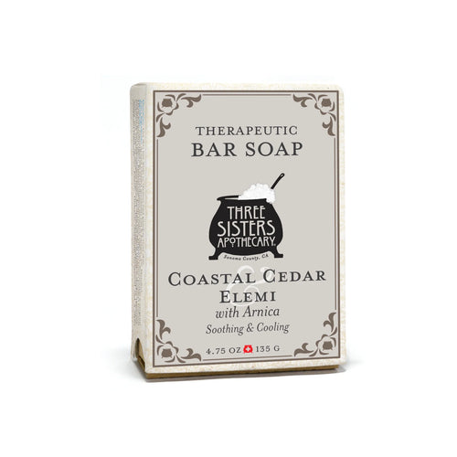 A 4.75 oz (135 g) Three Sisters Apothecary Coastal Cedar & Elemi Therapeutic Bar Soap in beige packaging with decorative borders and a black cauldron graphic, featuring Arnica for added benefit.