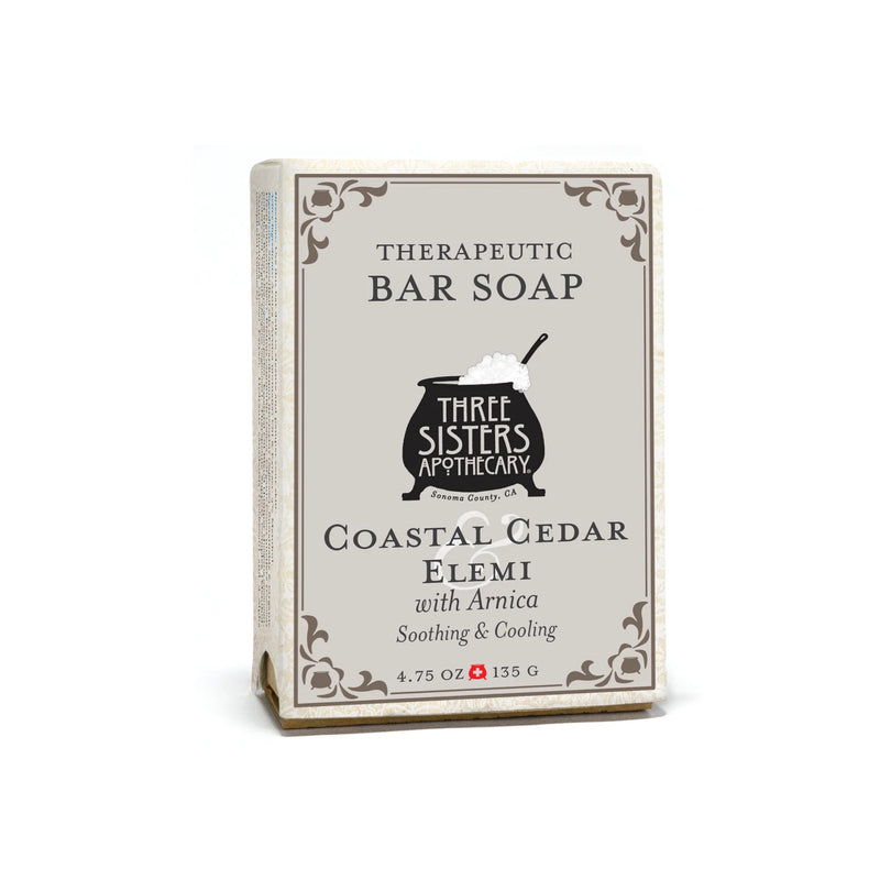 A 4.75 oz (135 g) Three Sisters Apothecary Coastal Cedar & Elemi Therapeutic Bar Soap in beige packaging with decorative borders and a black cauldron graphic, featuring Arnica for added benefit.