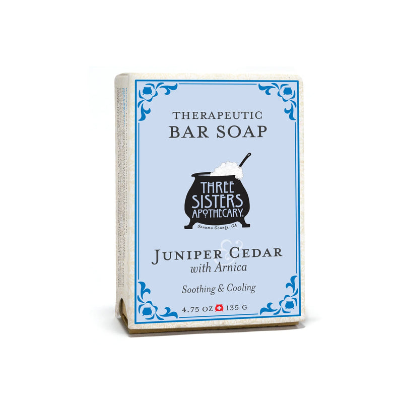 A package of Three Sisters Apothecary Juniper & Cedar Therapeutic Bar Soap (4.75 oz/135 g) features a black cauldron illustration on a light blue background, highlighting its soothing and cooling properties.