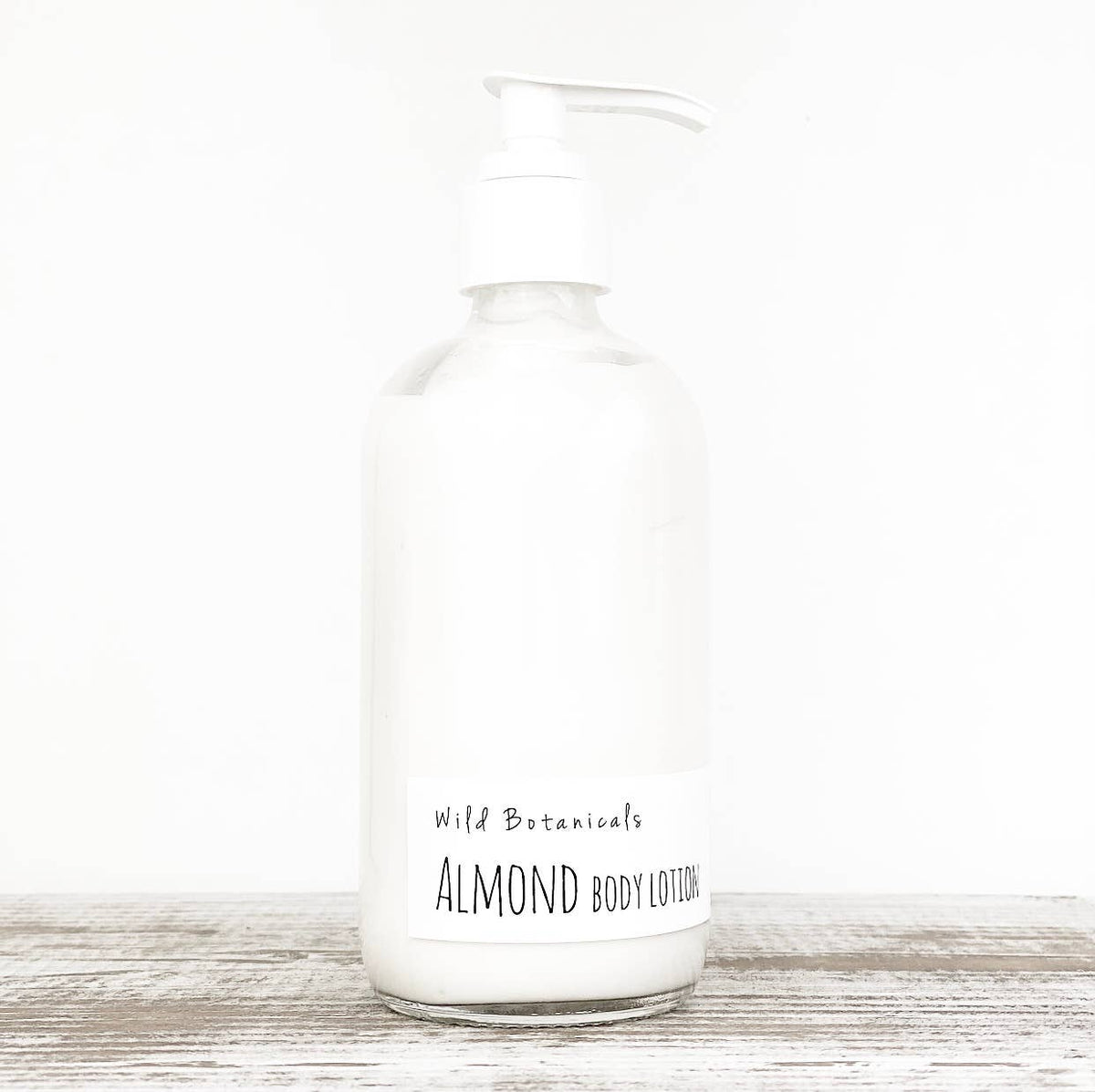 A clear glass pump bottle filled with white almond lotion from Wild Botanicals, enhanced with kokum butter, is set against a plain white background. The label on the bottle reads "Wild Botanicals Almond Lotion" in black text.