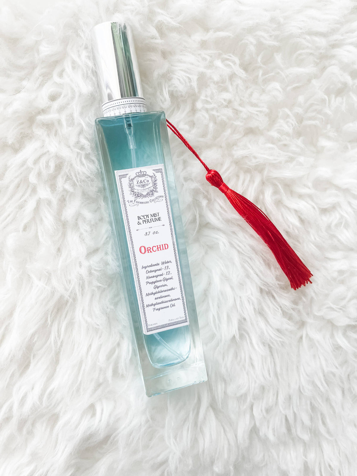 A clear glass bottle labeled “Z&Co. Farmhouse Body Mist - Orchid” with a silver cap and red tassel rests on a soft white surface, showcasing light blue, exotically scented mist and elegant decorative details.