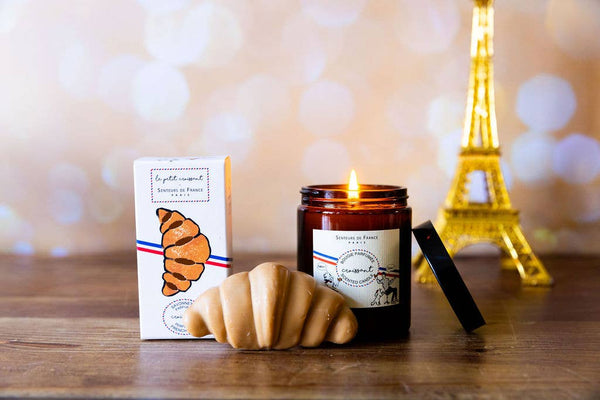 The Senteurs De France "Croissant" candle in a brown jar, croissant-shaped soap, and a box featuring a croissant illustration sit on wood, with an Eiffel Tower figurine adding Parisian décor flair in the background.
