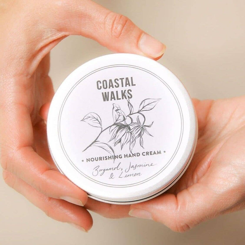 Close-up of hands holding a round tin of "Norfolk Natural Living Coastal Walks Hand Cream - 100ml" with a minimalist illustration of flowers on the lid. The label reads "Bergamot, Jasmine & Lemon with herbal extracts." The background is light and neutral, emphasizing its moisturizing properties.