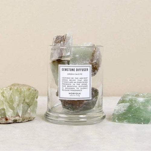 The Norfolk Natural Living Gemstone Green Calcit Diffuser features large green and brown calcite stones in a glass container, ideal for aromatherapy oils, displayed on a light surface with two extra stones and a beige background.