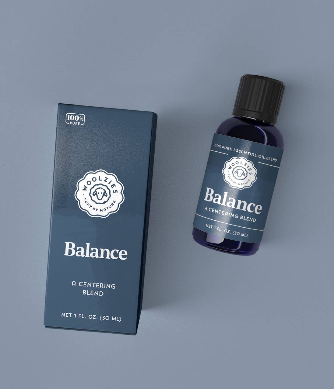 A blue bottle of Woolzies Balance Essential Oil Blend 1oz. and its matching box, both showing the Woolzies logo, rest on a gray surface. The label notes it's a grounding, 100% pure essential oil (1 fl. oz./30 ml).