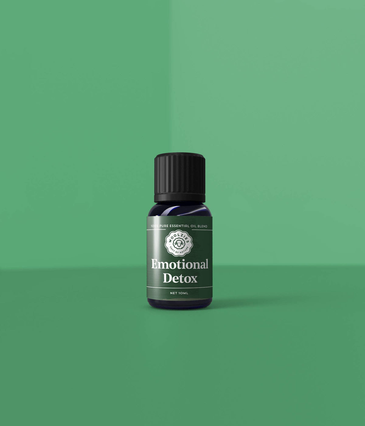 A small dark bottle of Woolzies Emotional Detox Essential Oil Blend stands upright on a smooth green surface, with a matching green background and soft lighting highlighting this Woolzies product.