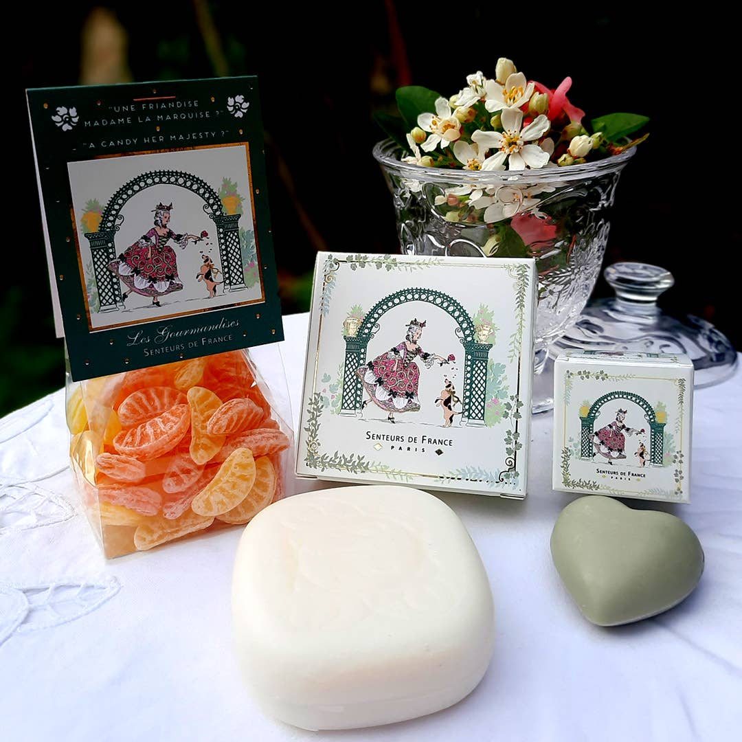 A display with French candies, a floral arrangement, decorative box and tin with vintage art, and Senteurs De France Olive Heart Soap—Marquise and Her Dog—celebrating French heritage on a white tablecloth.