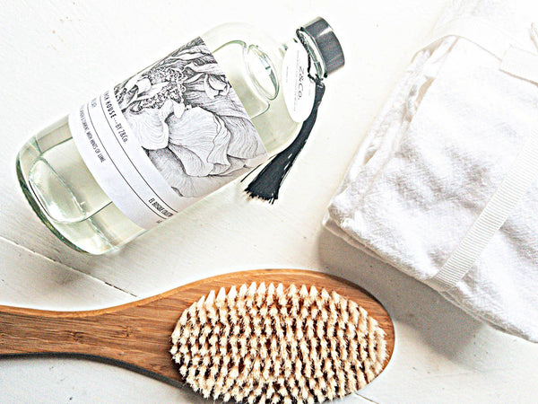 A Z&Co. French House El Bosque Bubble Bath bottle, featuring a floral label and black tassel, sits beside a folded white towel and a wooden body brush with light bristles, elegantly placed on a pristine white surface.