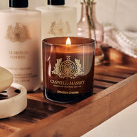 A Caswell-Massey Candle - Golden Embers sits on a wooden bath tray beside almond lotion and a small vase, its soy-blend meditative fragrance filling the air for a cozy, relaxing atmosphere.