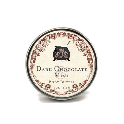 A tin container of Three Sisters Apothecary Dark Chocolate & Mint Body Butter enriched with natural botanicals. The elegantly decorated label highlights the product's weight of 4 oz. (113 g) and prominently displays the Three Sisters Apothecary logo at the top center.