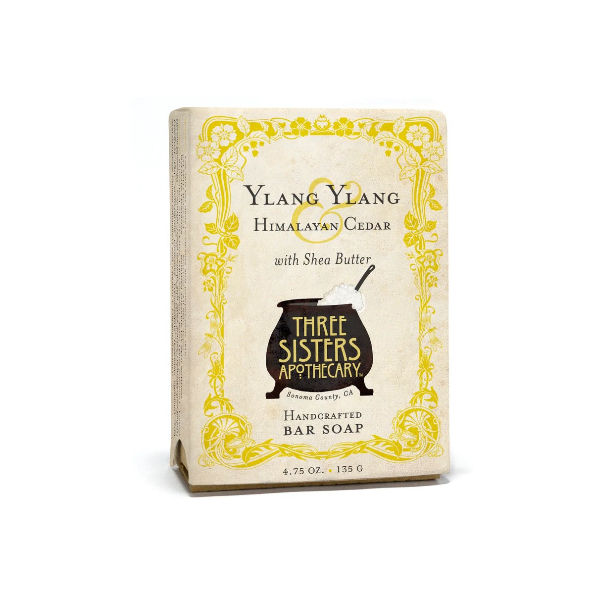Three Sisters Apothecary Ylang Ylang & Cedar Bar Soap comes in a cream box with yellow floral accents, features a black cauldron logo, and is handcrafted to nourish skin. Each bar weighs 4.75 oz.