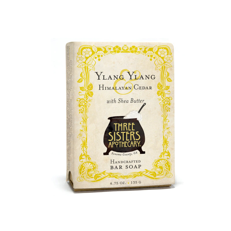 Three Sisters Apothecary Ylang Ylang & Cedar Bar Soap comes in a cream box with yellow floral accents, features a black cauldron logo, and is handcrafted to nourish skin. Each bar weighs 4.75 oz.