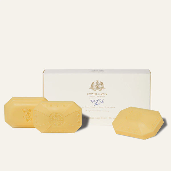 The Caswell-Massey Elixir of Love Three-Soap Set includes three hexagonal, plant-based triple-milled soaps with decorative embossing, displayed before a white Caswell-Massey box accented in gold and blue with the brand logo and product details.